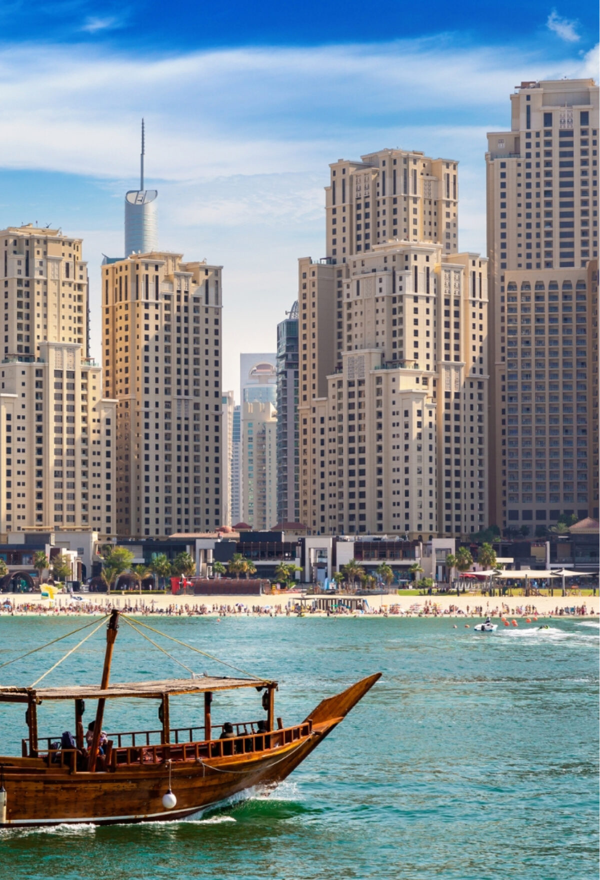 A traditional wooden dhow sailing across turquoise waters with Dubai's iconic beachfront skyscrapers in the background under a bright blue sky.