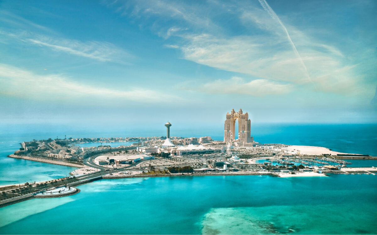 Aerial view of an island with turquoise waters, featuring iconic buildings, including a luxurious hotel and a tall observation tower, surrounded by marinas and sandy beaches under a bright blue sky.