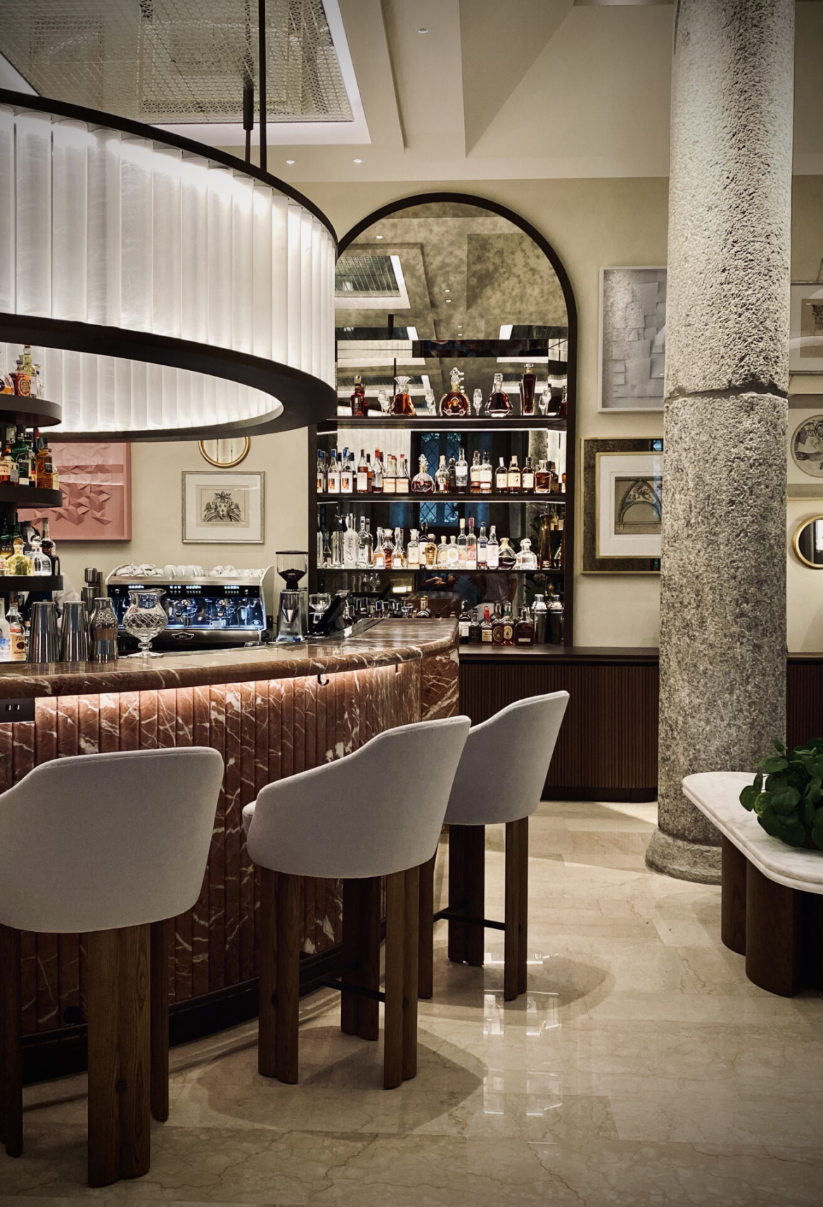 A sophisticated bar with a marble counter, plush stools, and a beautifully illuminated design, featuring an arched mirror and shelves stocked with premium spirits.