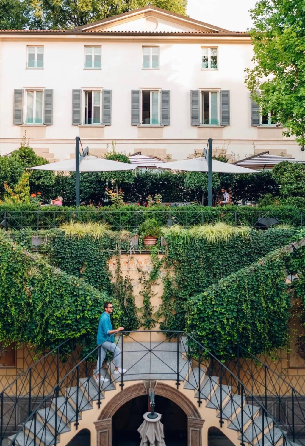 A charming multi-level garden adorned with lush greenery and ivy-covered walls, leading to a historic villa with elegant windows and shaded outdoor seating.